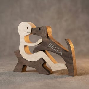 May include: A wooden sculpture of a person and a dog embracing. The person is light-colored, and the dog is dark brown with the name "BELLA" engraved on its side. The sculpture is made of wood and has a minimalist design.