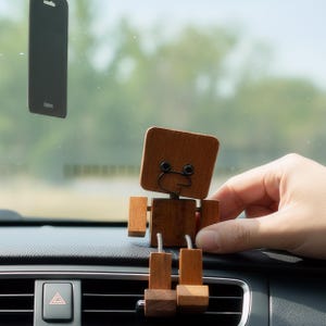 Puede incluir: Una figura de robot de madera con una cabeza cuadrada y partes del cuerpo en forma de bloque. El robot tiene brazos de alambre y una cara simple. Está sentado en el salpicadero de un coche. Un objeto rectangular negro cuelga en el fondo.