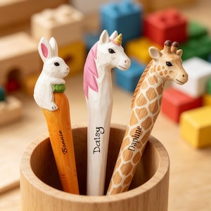 Peut inclure: Trois stylos décoratifs dans une tasse en bois. Les stylos présentent des motifs d'animaux : un lapin blanc sur une carotte, une licorne blanche avec une crinière rose et une girafe. Le stylo lapin est étiqueté "Bonnie", le stylo licorne est étiqueté "Daisy", et le stylo girafe est étiqueté "Daphne".
