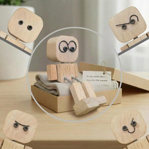 Peut inclure: Une figurine articulée en bois avec une tête et un corps carrés, des yeux en fil de fer noir et une expression neutre. La figurine est dans une boîte cadeau avec une carte qui dit : "To [Recipient's Name], Enjoy the little moments!"