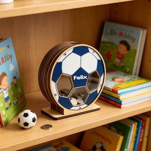 以下が含まれることがあります： 青と白のデザインの木製サッカーボール型貯金箱。名前「Felix」が見えます。小さなサッカーボールと本が棚にあります。貯金箱にはコインを挿入するためのスロットと、中のコインを見るための透明な窓があります。