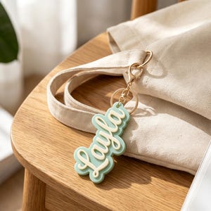 May include: A custom keyring featuring the name Layla in a raised, cream-coloured script font set against a scalloped, sage green background. The charm is attached to a gold-toned metal lobster clasp and a circular split ring for easy attachment.