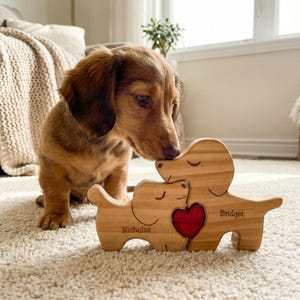 May include: A wooden dachshund dog puzzle with a red heart in the center, personalized with the names "Nicholas" and "Bridget". A brown dachshund puppy is looking at the puzzle. The puzzle is on a cream-colored carpet.