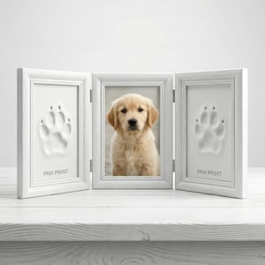 May include: A white, hinged picture frame with three panels. The center panel holds a photo of a golden retriever puppy. The side panels feature paw print impressions with the text "PAW PRINT" below each. The frame is on a white surface.
