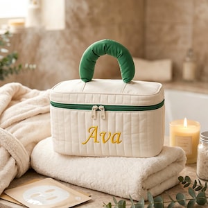 May include: A cream-colored quilted vanity case with a green handle and green zipper trim. The name Ava is embroidered in yellow script on the front. The rectangular bag features vertical stitching and two silver-toned zipper pulls.