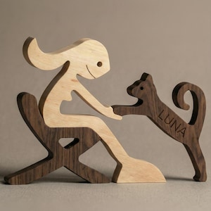 May include: A wooden sculpture depicting a person seated in a chair, interacting with a cat. The person is light-colored wood, while the chair and cat are dark wood. The cat's name, "LUNA", is carved into its side. The sculpture is a decorative item.