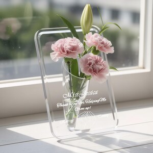 May include: A clear acrylic vase with a rectangular frame, holding pink carnations and a lily bud. The vase is engraved with the text "Miss Jessica, Thank You For Helping Me Grow up!" and names. A thoughtful gift.
