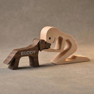 Puede incluir: Escultura de madera de una persona arrodillada saludando a un perro. El perro es marrón con el nombre "BUDDY" grabado en su costado. La persona es de madera clara y el perro es de un marrón más oscuro. La escultura está sobre un fondo neutro.