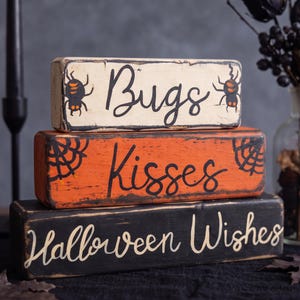 Puede incluir: Una pila de tres bloques rectangulares de madera con bordes desgastados. El bloque superior es crema con arañas negras y el texto Bugs. El bloque central es naranja con telarañas negras y el texto Kisses. El bloque inferior es negro con el texto Halloween Wishes.