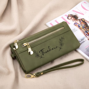 May include: Olive green wallet with gold-tone zippers and hardware. The wallet features the name "Theodora" in script with floral accents. Includes a wrist strap and is displayed on a neutral surface.