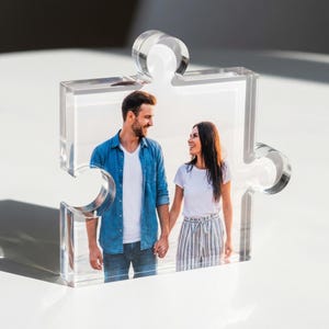 May include: Clear acrylic puzzle piece photo frame. The frame holds a colour photograph of a couple holding hands. The frame is transparent and has a glossy finish. The background is blurred.