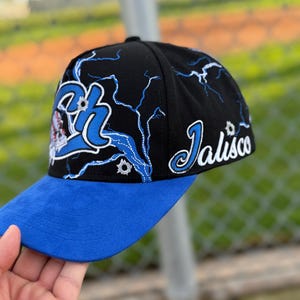 May include: Black baseball cap with a blue brim. The cap features the word "Jalisco" in white script on the side and a blue and white design on the front. Lightning bolt graphics are also present.