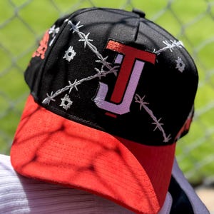 May include: Black baseball cap with a red brim. The cap features a white barbed wire design and a stylized "J" and "T" logo in red and white. The cap is resting on a white and gray surface.