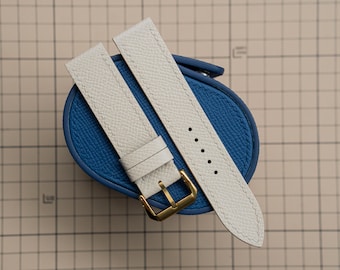 Peut inclure: Deux bracelets de montre en cuir blanc cassé avec des boucles dorées, posés sur un étui en cuir bleu. Les bracelets ont une surface texturée et des bords cousus. Le fond est un quadrillage.