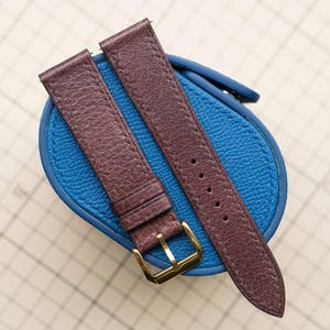 May include: Two brown leather watch straps with a gold-colored buckle, resting on a blue leather surface. The straps have a textured appearance and are designed for a watch. The background is a light grid pattern.