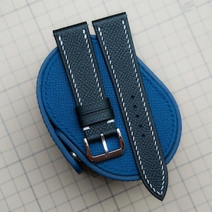 Puede incluir: Dos correas de reloj azul marino con costuras blancas y una hebilla plateada, sobre un estuche de cuero azul. Las correas tienen una superficie texturizada.