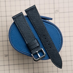 May include: Two navy blue leather watch straps with silver buckles on a blue leather case. The straps have a textured surface and are designed for a watch. The case is round and a matching blue colour.