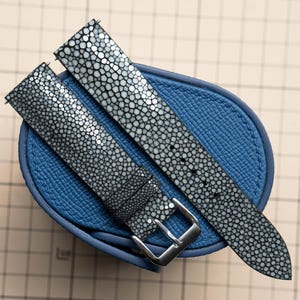 May include: A two-piece watch strap featuring a textured, grey and white stingray skin pattern. The strap includes a polished silver-tone metal buckle and is positioned over a small, oval-shaped blue leather case with a pebbled surface texture.