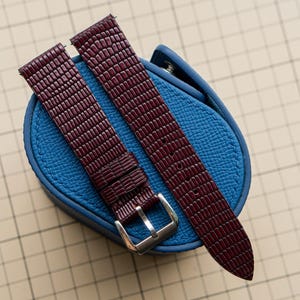 Peut inclure: Un bracelet de montre bordeaux foncé avec un motif texturé façon lézard et une boucle en métal argenté, posé sur un petit étui de rangement rond bleu vif texturé avec un bouton de fermeture à pression visible.