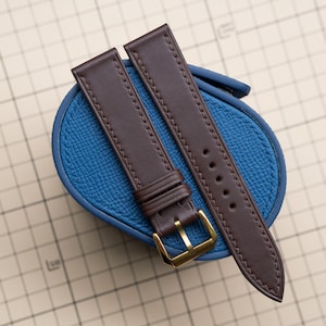 Puede incluir: Dos correas de reloj de cuero marrón con hebillas y costuras doradas, apoyadas sobre una superficie de cuero azul. Las correas son nuevas y sin usar, mostrando un diseño clásico. Las correas son una pieza de repuesto.