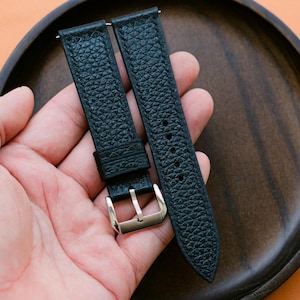 Puede incluir: Correas de reloj de cuero negro con superficie texturizada y hebilla plateada. Las correas se muestran en una bandeja de madera, mostrando su diseño. Las correas miden aproximadamente 23 cm de largo.