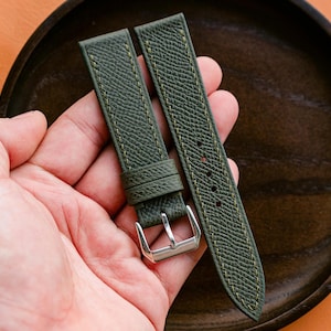 Op de afbeelding: Olijfgroene horlogeband met een getextureerd oppervlak en contrasterende gele stiksels. De band heeft een zilveren gesp en wordt getoond op een donkerhouten bord tegen een oranje achtergrond. Een stijlvol accessoire.