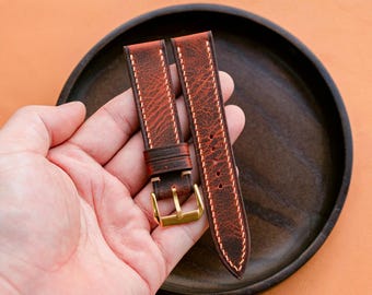 May include: A pair of brown leather watch straps with white stitching and gold-colored buckles. The straps are held in a hand, resting on a dark wooden plate against an orange background. The leather has a distressed appearance.