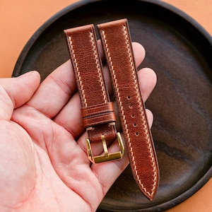 May include: Brown leather watch straps with white stitching and a gold-colored buckle. The watch bands are held in a hand, showcasing the detailed stitching and the quality of the leather. The background is a dark wooden plate on an orange surface.