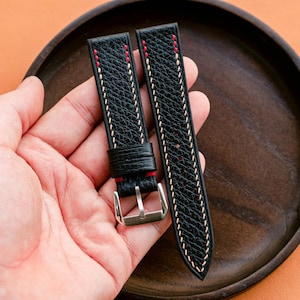 Può includere: Cinturini per orologi in pelle nera con cuciture bianche e rosse. I cinturini hanno una superficie strutturata e una fibbia argentata. I cinturini sono esposti su un vassoio di legno.