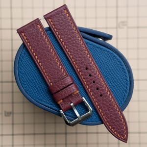 Puede incluir: Correas de reloj de cuero burdeos con costuras naranjas. Las correas son texturizadas y tienen una hebilla plateada. Las correas se muestran sobre una funda redonda azul.