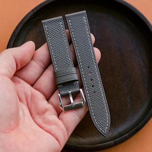 Puede incluir: Correa de reloj gris con costuras blancas y hebilla plateada. La correa tiene una superficie texturizada y un diseño clásico. La correa del reloj se muestra sobre un plato marrón oscuro sobre un fondo naranja.