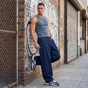 May include: A person wearing a gray tank top, dark blue jeans, and sneakers, leaning against a brick wall with graffiti. The jeans are loose-fitting. The sneakers are white, navy, and red.