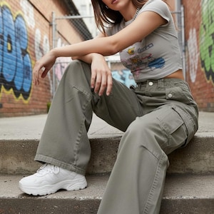 May include: A person wearing a gray crop top with a cartoon graphic, olive green cargo pants, and white sneakers. The person is sitting on concrete steps in front of a brick wall with graffiti.