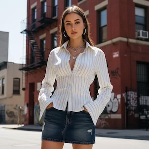 以下が含まれることがあります： 白いシャツに青い縦縞とフレア袖、濃い色のデニムミニスカートを合わせたコーディネート。モデルはシルバーのフープイヤリングと蝶のペンダントネックレスを着用しています。