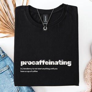 May include: Black t-shirt with the word "procaffeinating" in white, along with a definition. A silver necklace with a sunburst pendant rests on the shirt. Blue jeans and a white shoe are visible.