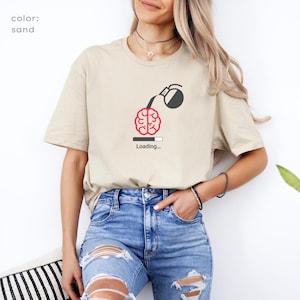 May include: Sand-coloured t-shirt with a graphic of a brain being filled with coffee from a coffee pot. The text "Loading..." is below the brain graphic. The model is wearing blue jeans.