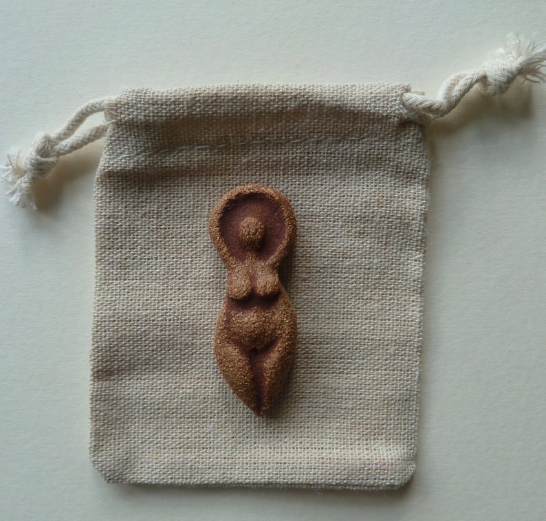 Puede incluir: Una peque&ntilde;a figurita de cer&aacute;mica marr&oacute;n de una mujer con la cabeza y los brazos levantados. La figurita est&aacute; sentada en una peque&ntilde;a bolsa de cord&oacute;n beige.