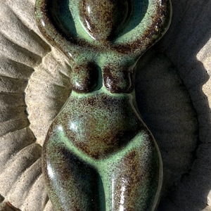 May include: A ceramic figurine of a stylised human form with arms raised above the head. The piece features a glossy glaze with a mottled colour palette of deep brown and light mint green, creating a smooth, reflective surface texture throughout.