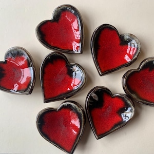 May include: Seven heart-shaped ceramic dishes. Each has a glossy red interior and a black and grey rim. The dishes are arranged in a circular pattern, highlighting their unique design and craftsmanship.