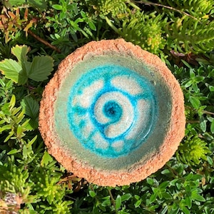 May include: A handmade ceramic bowl with a spiral design in shades of blue and white. The bowl has a textured, light brown rim and is surrounded by green foliage. The interior features a glossy finish.
