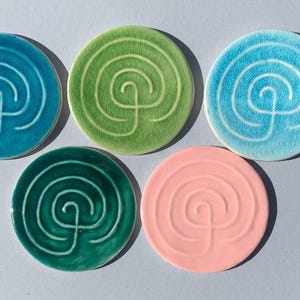 Peut inclure: Cinq dessous de verre ronds en céramique dans les tons de turquoise, vert et rose. Chaque dessous de verre présente un motif en spirale blanc. Les dessous de verre sont disposés sur une surface gris clair.