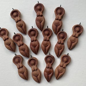 Puede incluir: Una colección de colgantes marrones con figuras femeninas estilizadas. Cada colgante es una representación detallada de una forma femenina, con una superficie texturizada y un pequeño lazo en la parte superior para colgar. Los colgantes están dispuestos sobre una superficie clara.
