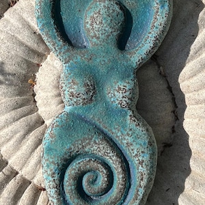 May include: A teal and dark blue textured wall plaque shaped like a stylised human figure with arms raised above the head and a prominent spiral design centred on the lower torso. The surface has a mottled, weathered appearance with dark brown accents.
