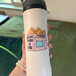 May include: White insulated water bottle with a black lid and handle. A sticker on the bottle features a pink pharmacy with flames and the text "EVERYTHING IS FINE".