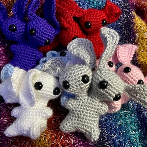 May include: A collection of crocheted bunny plushies in various colors, including purple, red, gray, white, and pink. The plushies are arranged on a colorful striped blanket.