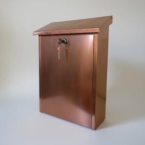 May include: A copper-colored mailbox with a slanted top and a key lock. The mailbox has a rectangular shape and a smooth, reflective surface. A key hangs in the lock. The mailbox is designed for outdoor use.