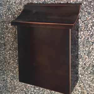 May include: A dark brown metal mailbox with a curved top and a rectangular opening. The mailbox is mounted on a wall of small stones.
