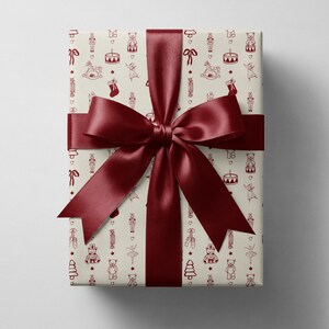 May include: A rectangular gift box wrapped in cream-colored paper featuring a repeating pattern of dark red line drawings of teddy bears, nutcrackers, drums, stockings, and ballet slippers, tied with a wide, dark red satin ribbon bow.