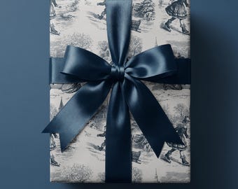 Puede incluir: Una caja de regalo rectangular envuelta en papel blanco roto con un patrón invernal de estilo toile en azul oscuro que muestra personas patinando sobre hielo y en trineo. El paquete está atado con una cinta de satén azul oscuro ancha.