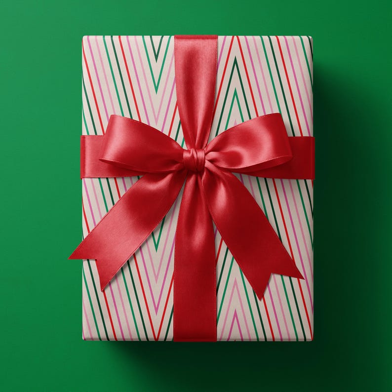May include: A rectangular gift box wrapped in cream-colored paper featuring a repeating pattern of thin vertical chevron stripes in red, green, and pink. The package is tied with a wide, smooth, deep red satin ribbon finished in a large bow.